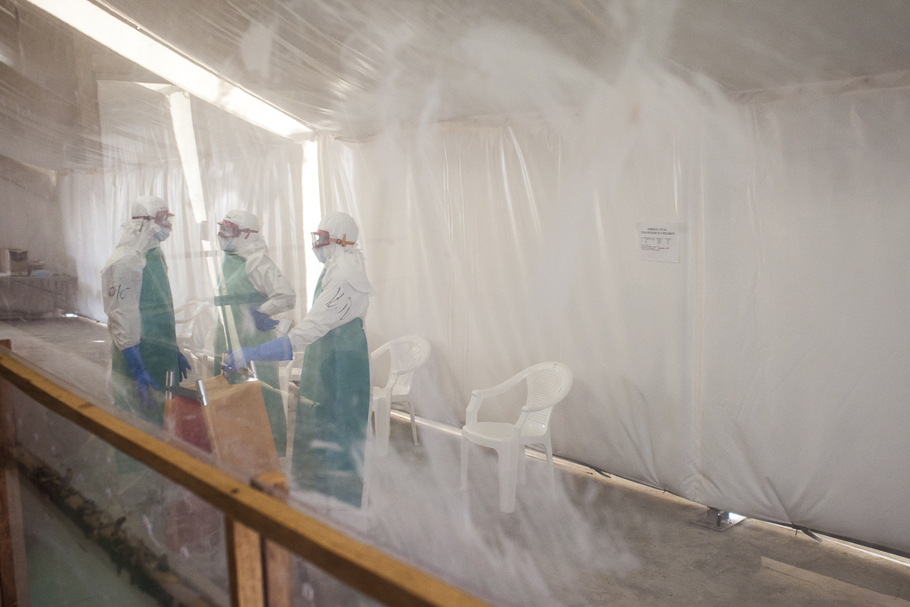 A view of the triage room in the high risk zone from a methacrylate spoiled by the chlorine solution used for disinfection. A team of hygienist with a folding stretcher is discussing their strategy to complete their task in the best possible way.  Communicate and move wearing the protective clothing is not an easy business, usually the hygienist teams draw up a plan together with the nurses before entering in the high risk zone. Ebola Treatment Center. Moyamba. Sierra Leone.